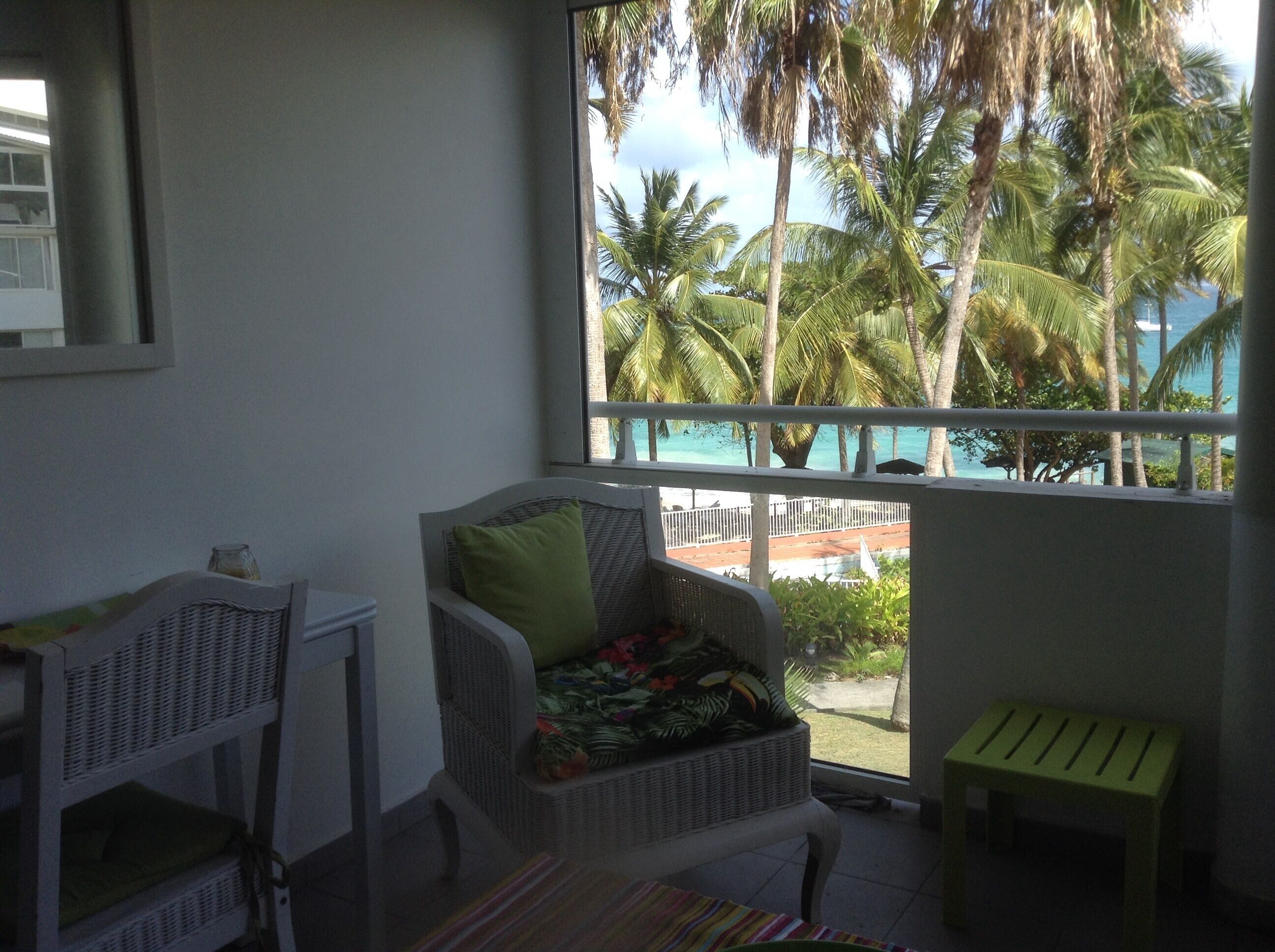 A room with a wicker chair, a small table, and a view of palm trees and a beach.
