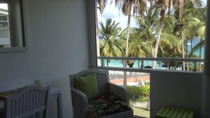 Quarto com cadeira de vime, uma mesa pequena e vista para palmeiras e uma praia.