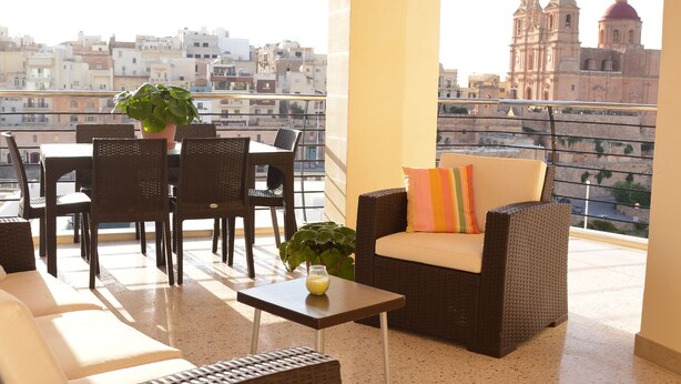 A balcony with wicker furniture, a potted plant, and a view of a cityscape including a cathedral. - Summer Haven, with breathtaking views and Private Swimming Pool (Mellieha)