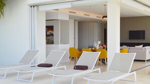 Un espace de détente extérieur moderne, avec des chaises longues blanches, un coin repas avec des chaises jaunes et un téléviseur à écran plat fixé au mur.