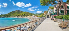 A coastal promenade with a beach, a stone bridge, and buildings with balconies overlooking the sea.