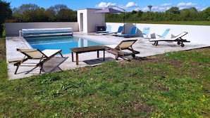 Ein Schwimmbecken mit Liegestühlen und einem Tisch, umgeben von einer Rasenfläche und Bäumen.