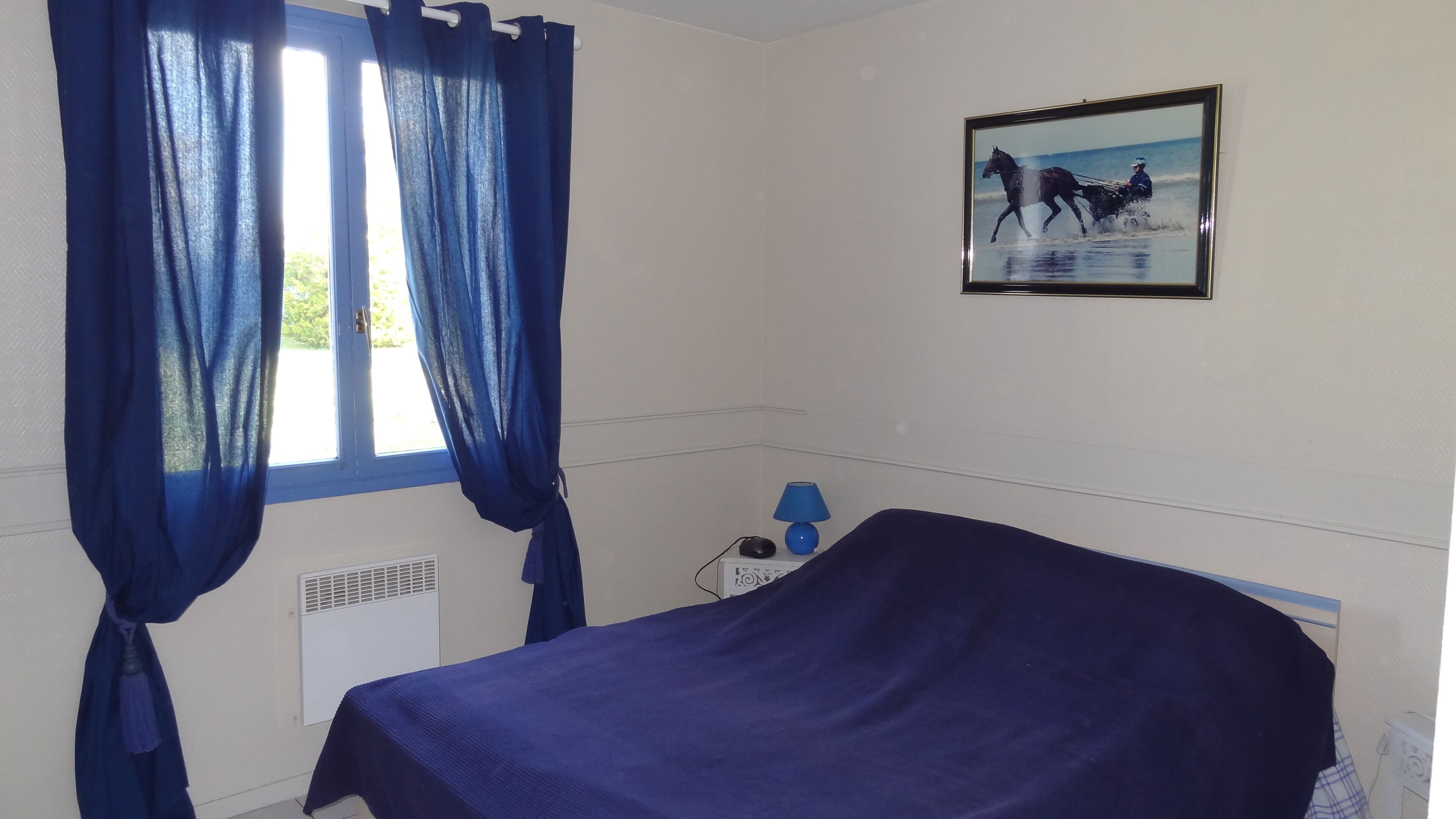 A room with a bed, a window with blue curtains, a wall-mounted picture of a horse and rider, and a small table with a lamp.