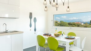 Une cuisine moderne avec un plan de travail blanc, un évier et un coin repas comprenant une table blanche et des chaises vertes. Des luminaires suspendus sont placés au-dessus de la table, et une fenêtre offre une vue sur la verdure et les montagnes.