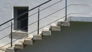 Un escalier avec des marches en pierre et une rampe métallique, menant à une fenêtre dotée d’un cadre blanc.