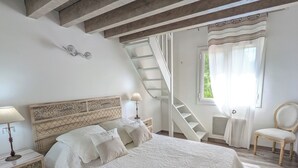 Ein Schlafzimmer mit einem großen Bett, einem Holz-Kopfteil, einem Nachttisch, einer Lampe, einem Sessel, einem Fenster mit Vorhängen und einer Treppe.