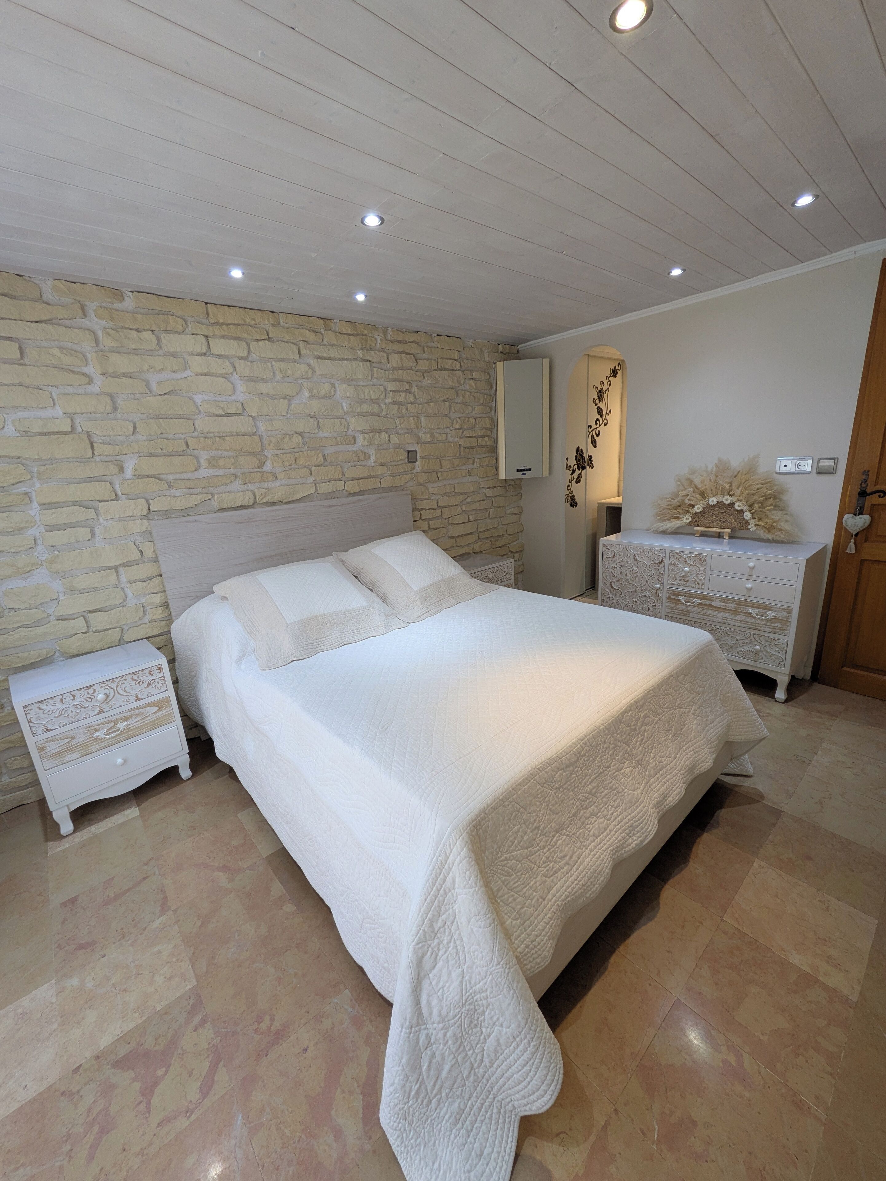 Una camera da letto con parete in pietra, un letto con copriletto, un comodino, una cassettiera e uno specchio.