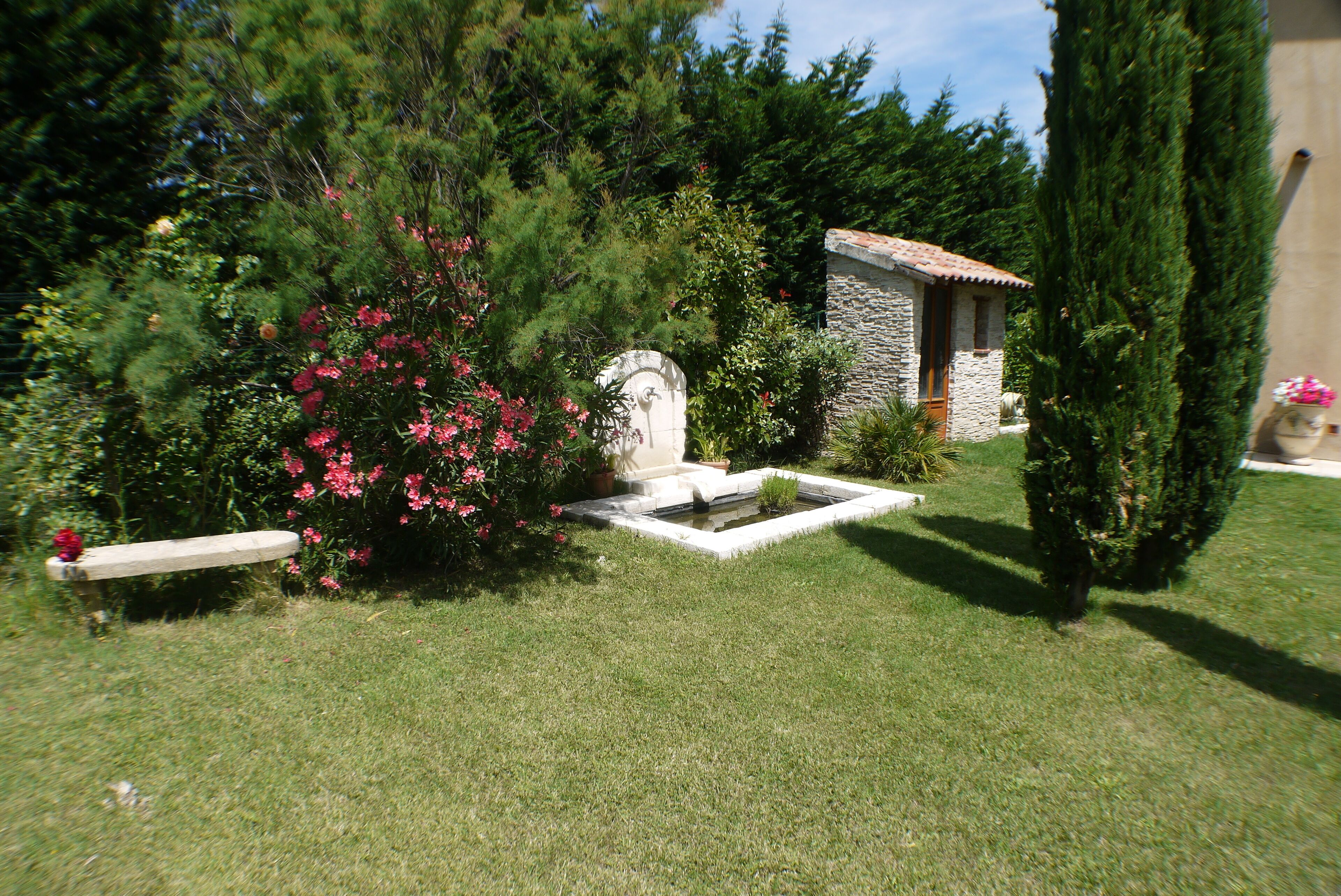 A stone building with a tiled roof, surrounded by lush greenery and a small fountain.