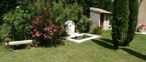 A stone building with a tiled roof, surrounded by lush greenery and a small fountain.