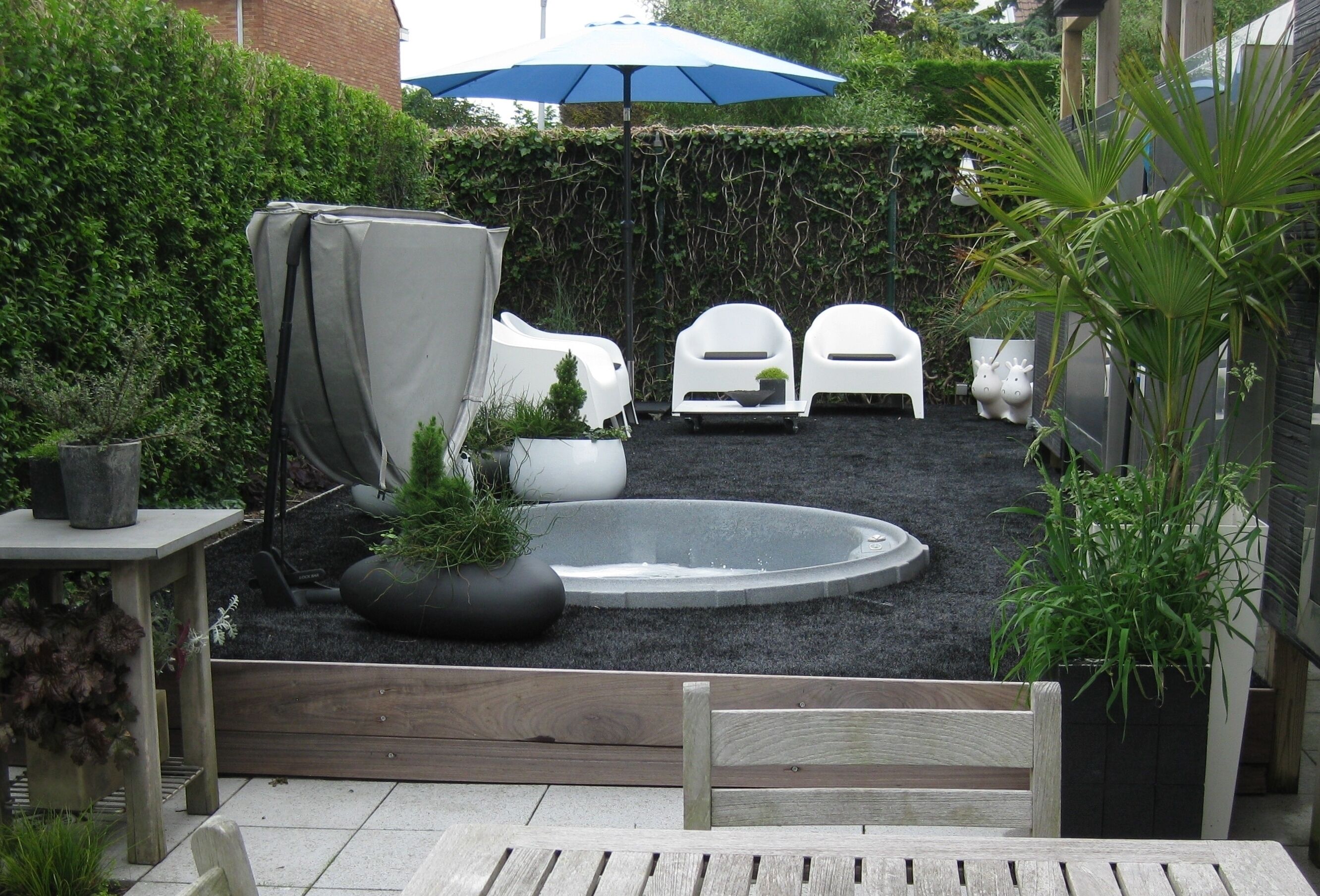 A backyard with a circular hot tub, white plastic chairs, a covered table, and various potted plants.