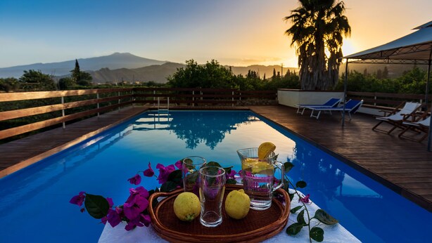 A poolside setting with a wooden deck, lounge chairs, and a table with drinks and flowers.