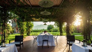 An outdoor dining area with a table set for six, surrounded by chairs, under a pergola with hanging lights.