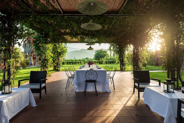 An outdoor dining area with a table set for six, surrounded by chairs, under a pergola with hanging lights.