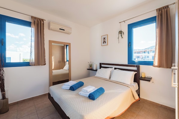A bedroom with a bed, a window, a mirror, a bedside table, and a view of buildings outside.