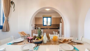 A dining area with a table set for two, a kitchen in the background, and a window with curtains. - Blue Sea Villa in Gennadi (Gennadi)