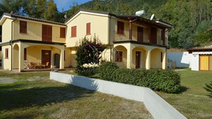 A two-story house with a balcony, a garden, and a hillside in the background.