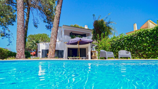A swimming pool with clear water, a covered seating area with chairs, and a building with a white facade and a red roof.