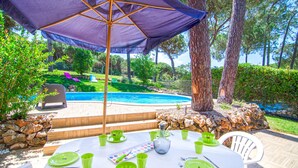 A poolside dining area with a table set for four, surrounded by greenery and a view of a swimming pool.