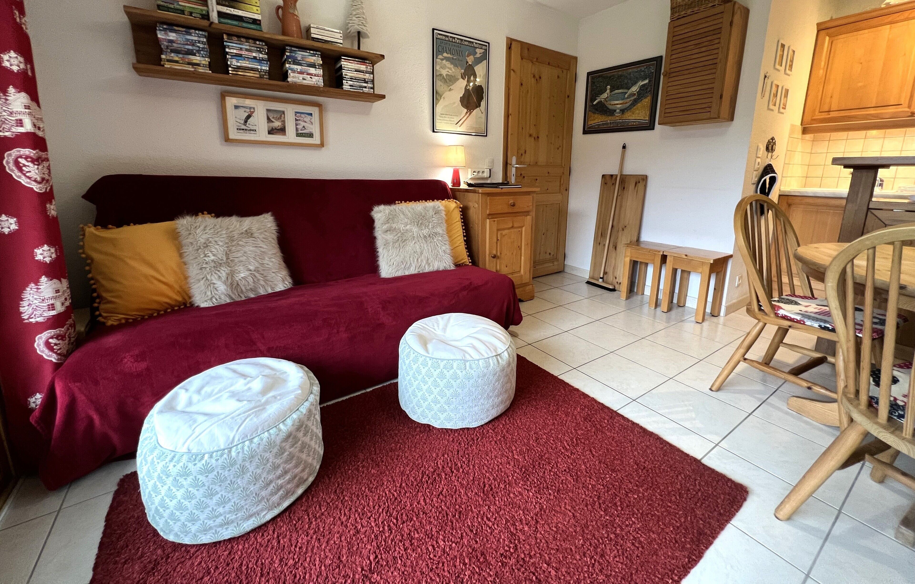 Een gezellige kamer met een rode bank, twee voetenbankjes, een houten eettafel en een keuken met houten kastjes en een betegelde spatwand.