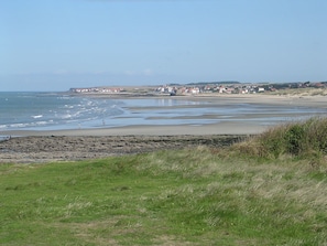 Een kustlandschap met een zandstrand, een grasveld op de voorgrond en een stad in de verte met huizen.