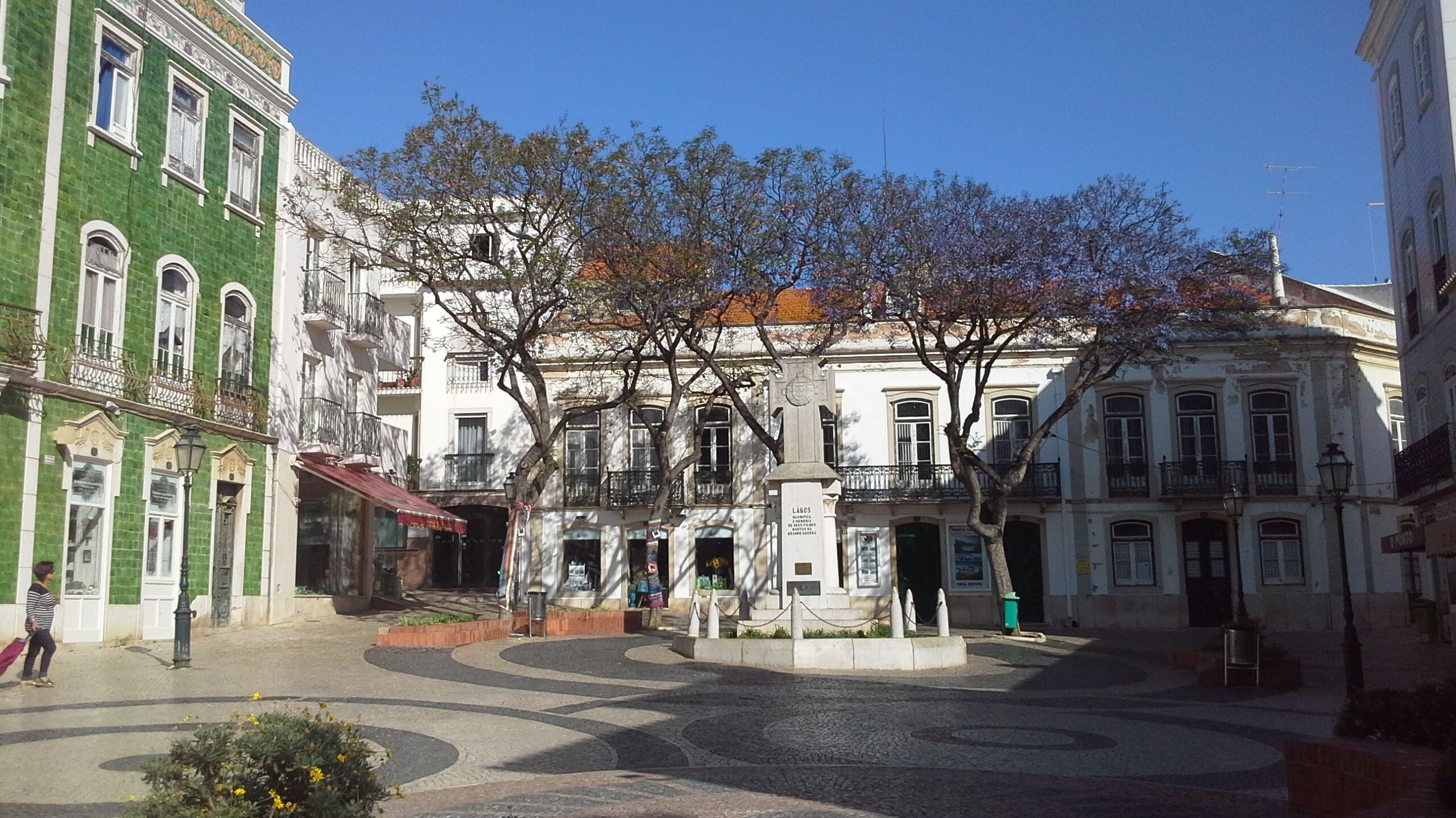 A historic square with a central monument, surrounded by classical architecture and a cobblestone pavement.
