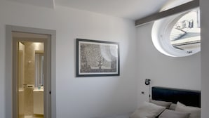 A bedroom with a bed, a round window, a framed picture, and a door leading to a bathroom. - Delightful two-room apartment in the heart of Genoa. (Genova)