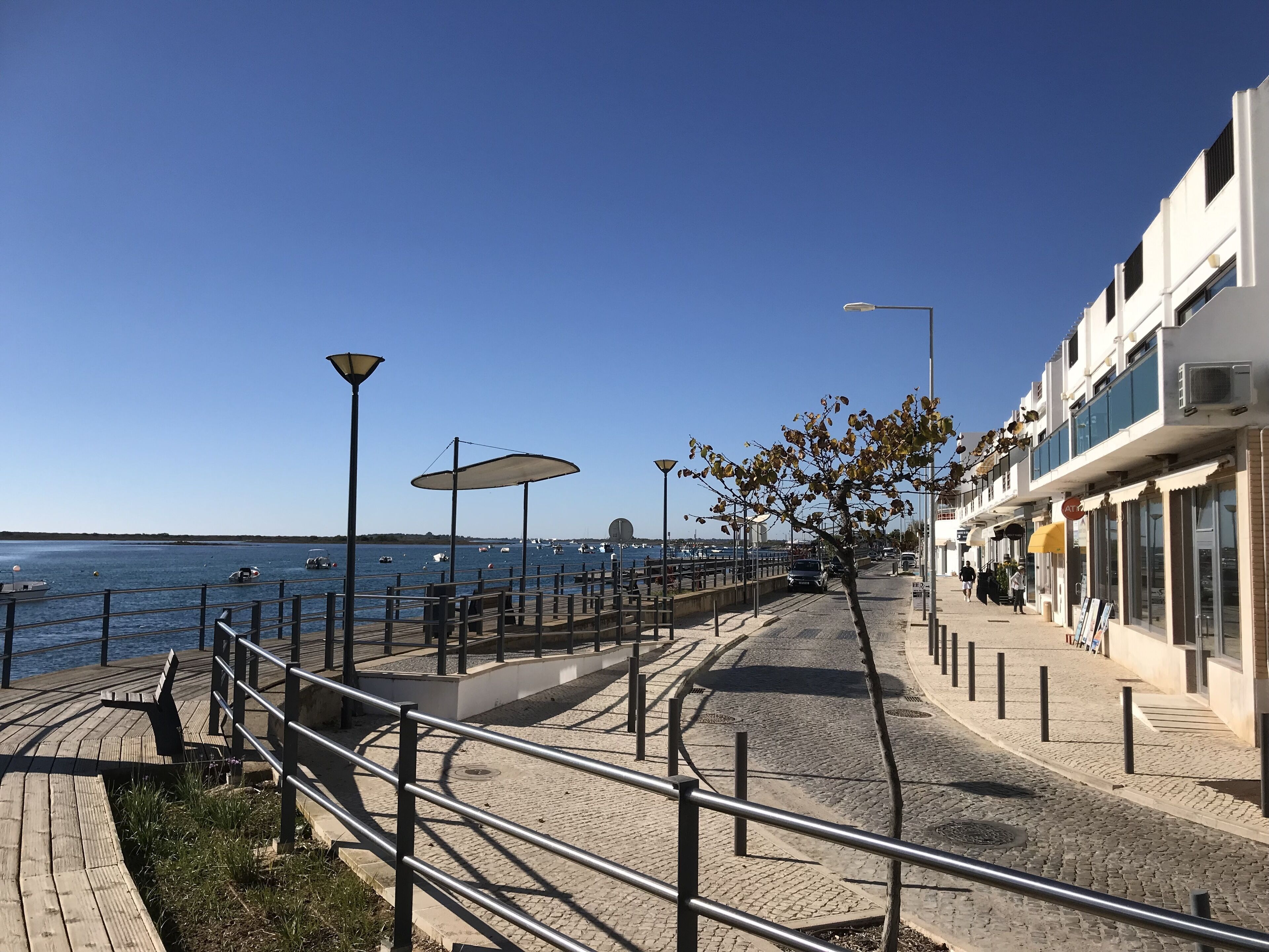 A waterfront promenade with a wooden walkway, metal railings, street lamps, and a row of shops.