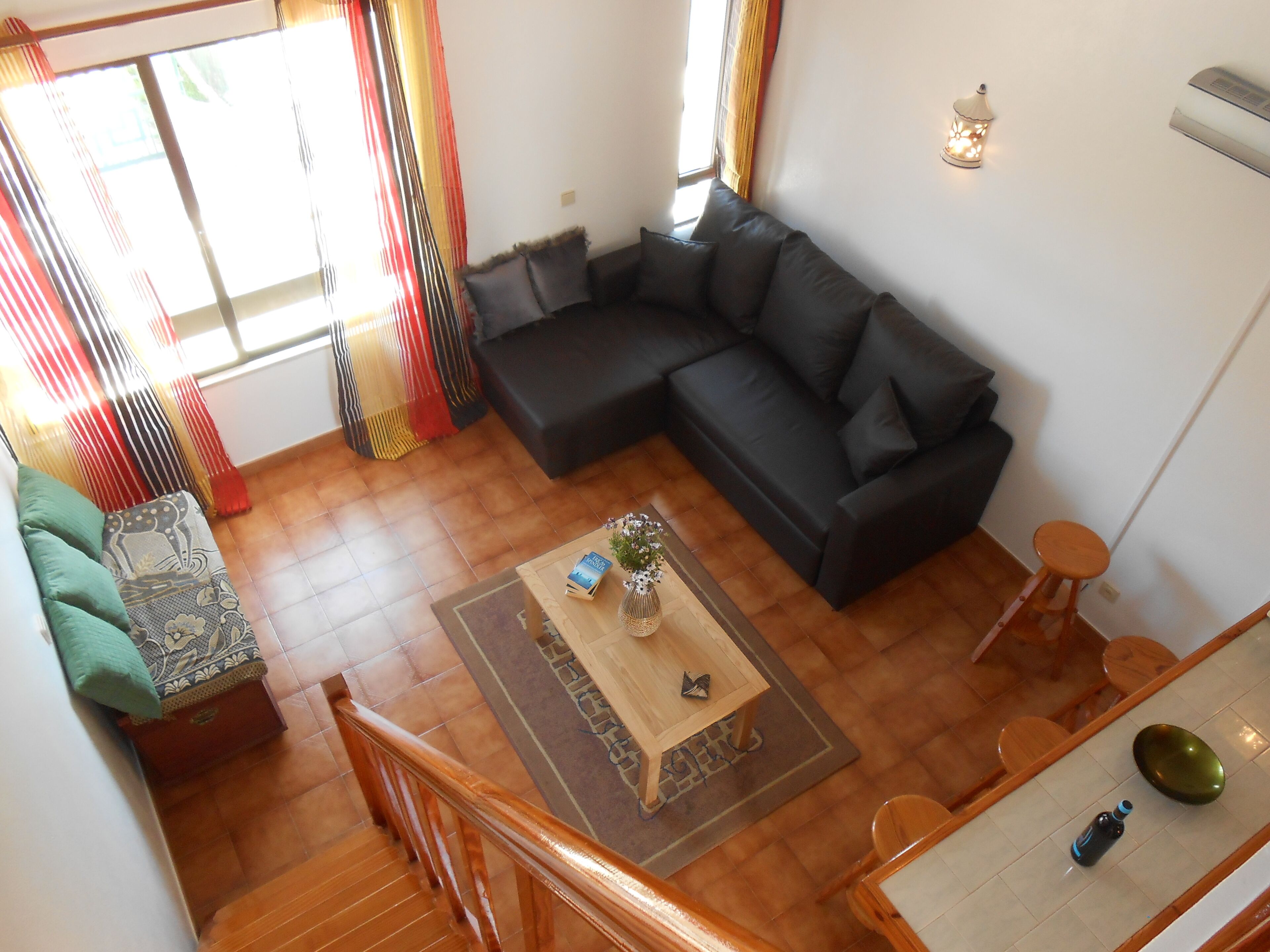 Un soggiorno con un divano modulare nero, un tavolino in legno con oggetti decorativi, una zona pranzo con tavolo e sedie in legno, e una scala con ringhiere in legno.