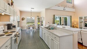 A modern kitchen with white cabinetry, a large island, and a dining area with a table and chairs. - Stunning 4 Bed Villa With Uninterupted Lake Views In The Sanctuary  (Davenport)