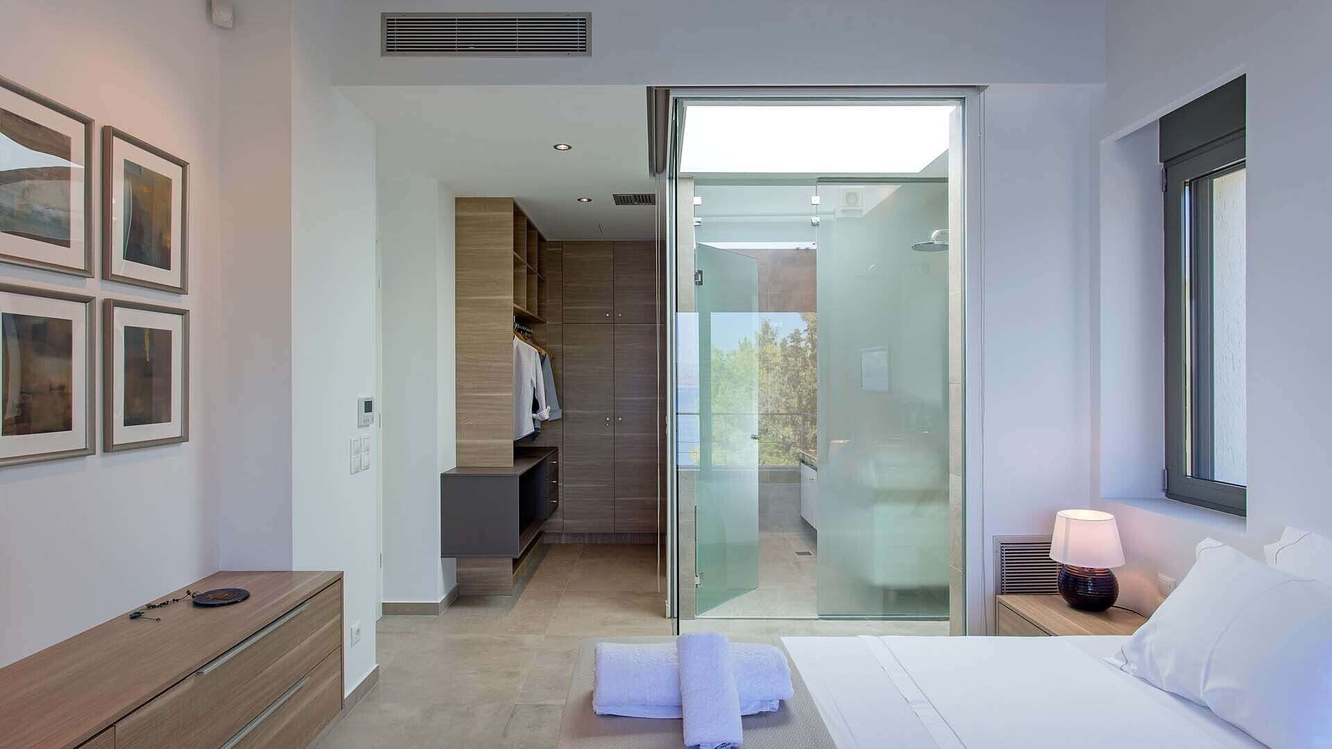 Une chambre à coucher moderne avec une porte en verre donnant sur une salle de bain, une table de chevet avec une lampe et des tableaux encadrés accrochés au mur.