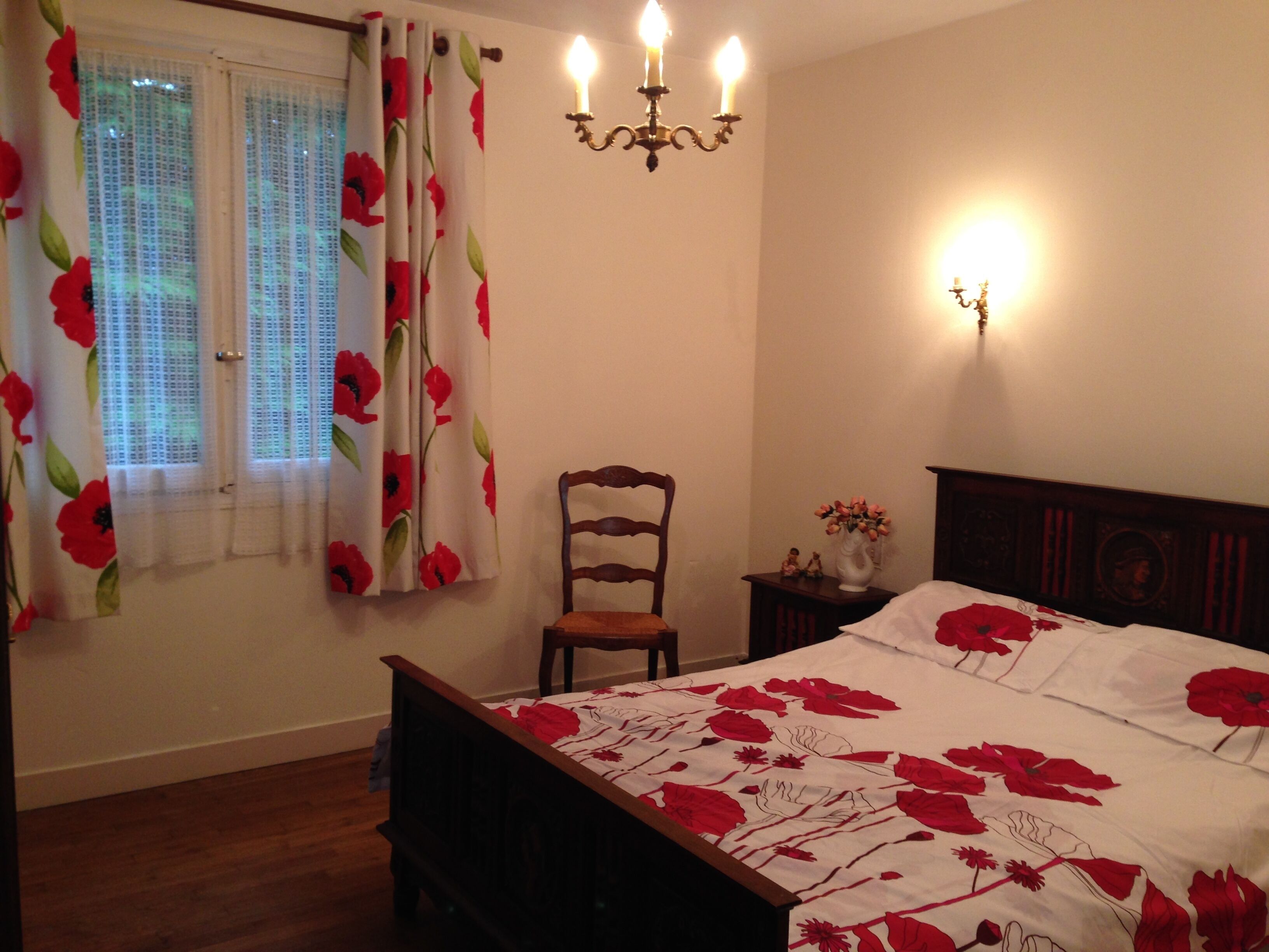 Un dormitorio con una cama de madera, una mesita de noche con un florero, una silla y una ventana con cortinas florales.