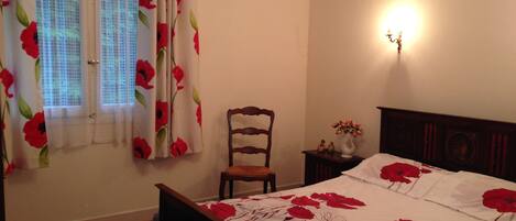 A bedroom with a wooden bed, a nightstand with a vase, a chair, and a window with floral curtains.
