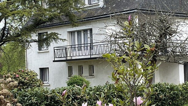 Una casa a due piani con un balcone, immersa nel verde e circondata da piante fiorite.