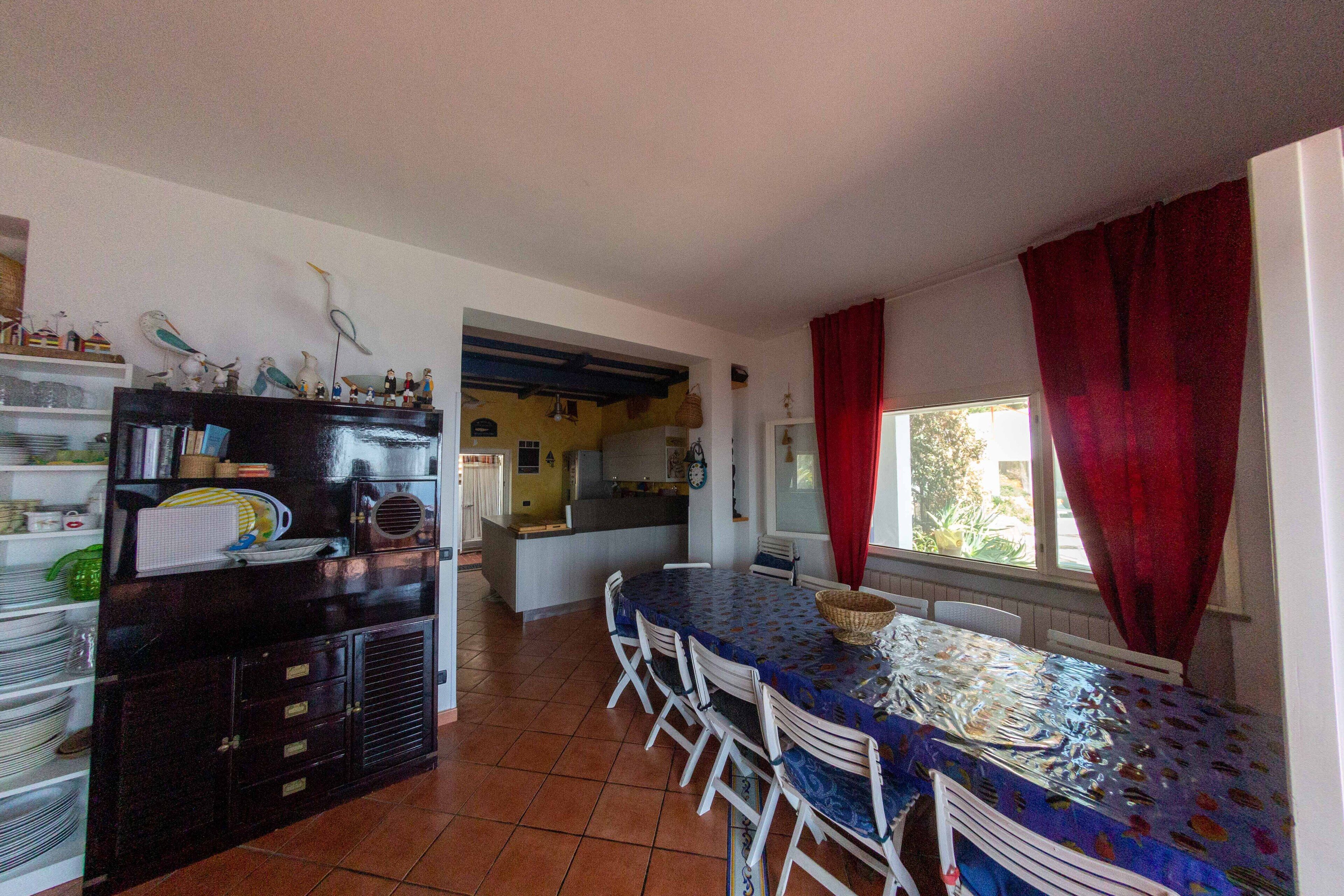 Un comedor con una mesa y sillas, una cocina al fondo y una ventana con cortinas rojas.