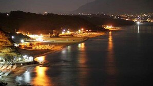 A nighttime coastal scene with a river, illuminated buildings, and a distant cityscape.