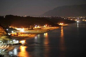 A nighttime coastal scene with a river, illuminated buildings, and a distant cityscape.