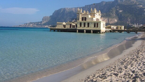 A coastal building with a prominent tower, situated on a sandy beach with clear blue waters and a mountain in the background.