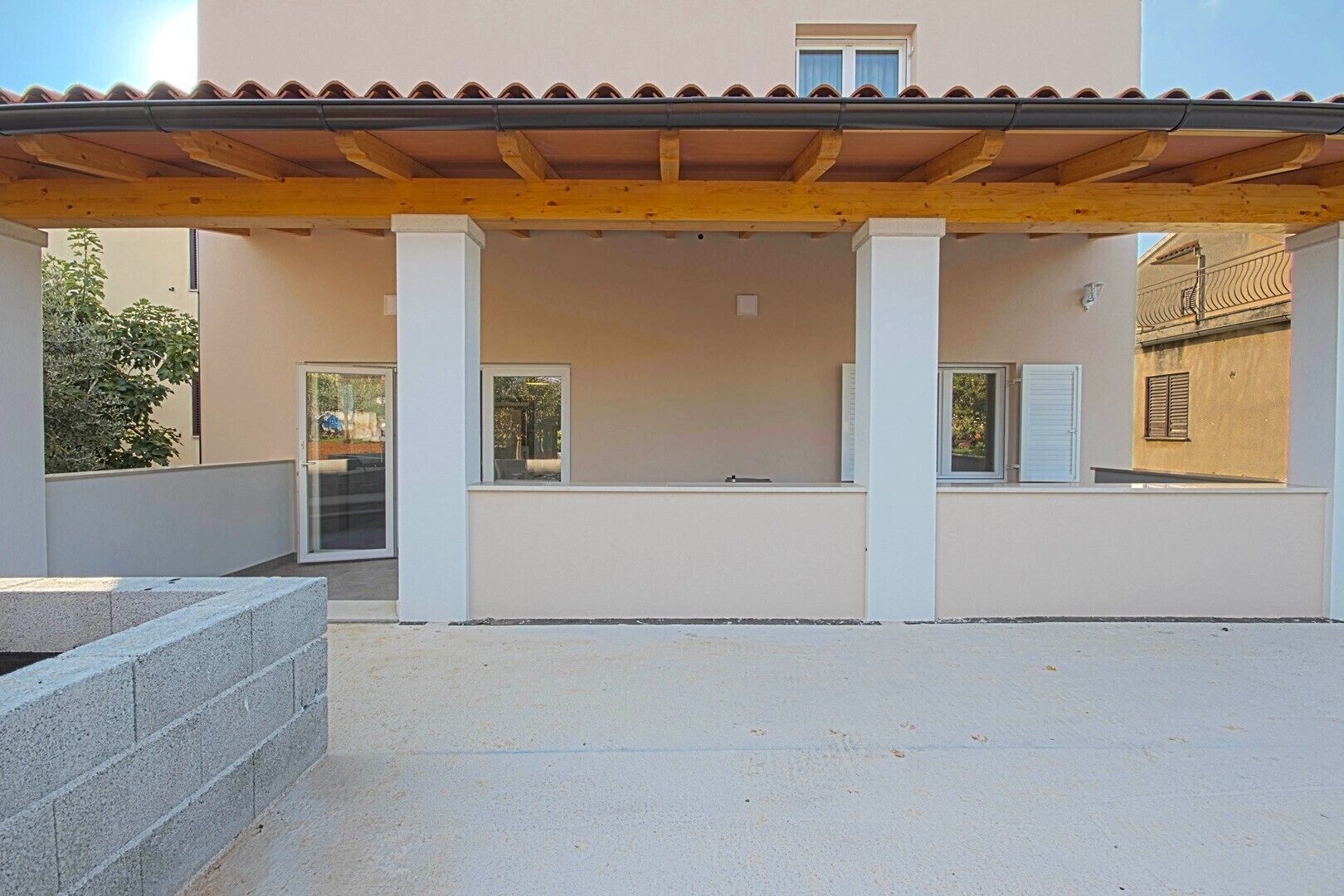 A covered outdoor area with a wooden pergola, a concrete bench, and a building with a tiled roof and windows.