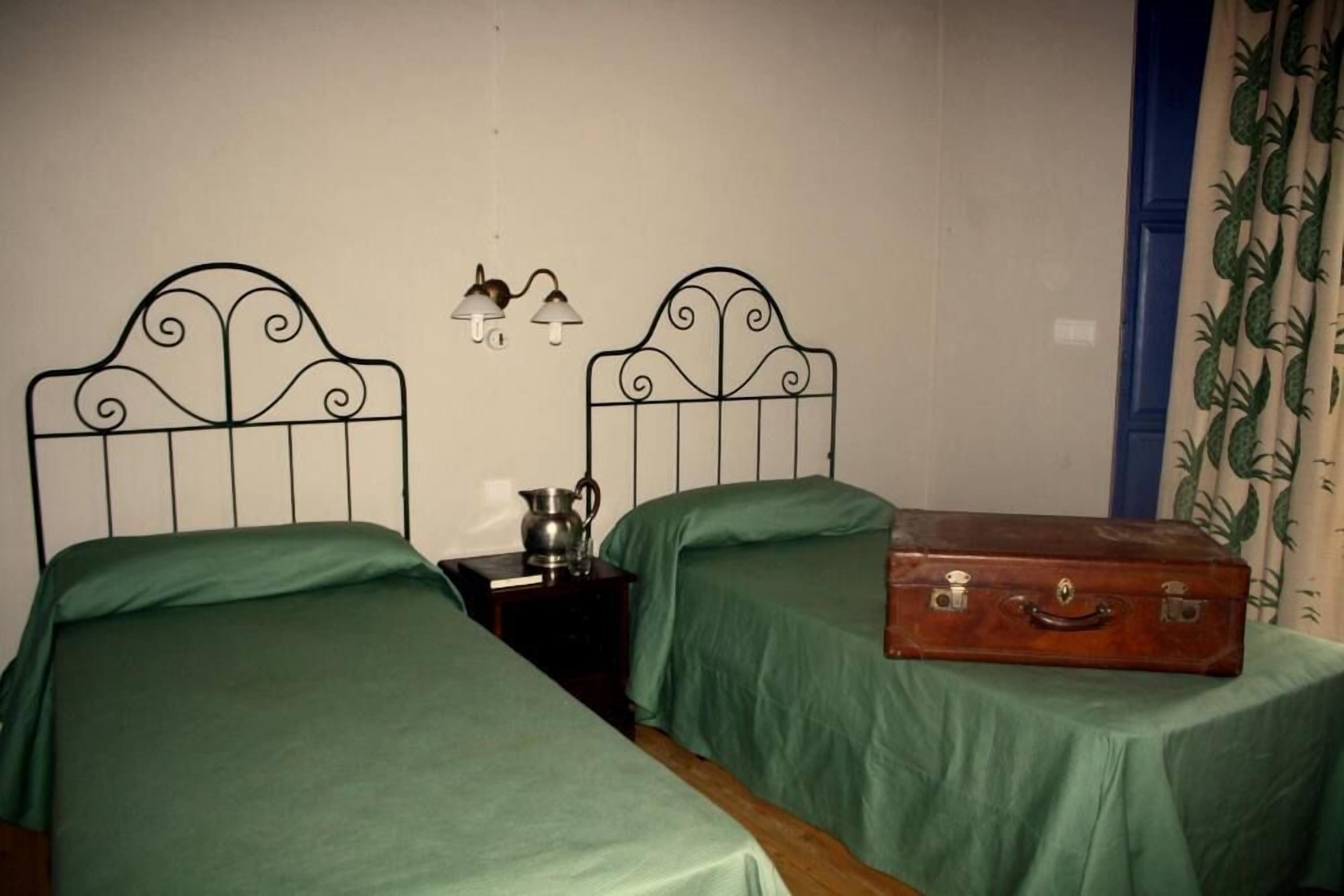 A room with two single beds with metal headboards, a nightstand with a lamp, and a suitcase on a table.