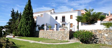 A white building with a stone wall, a pathway, and a large tree.
