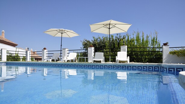 Une piscine entourée d’une clôture et d’arbres, avec des chaises longues blanches et des parasols.