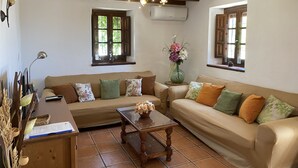 Un salon chaleureux avec un canapé, une table basse et un vase rempli de fleurs. Il y a des fenêtres avec des volets et un ventilateur de plafond.
