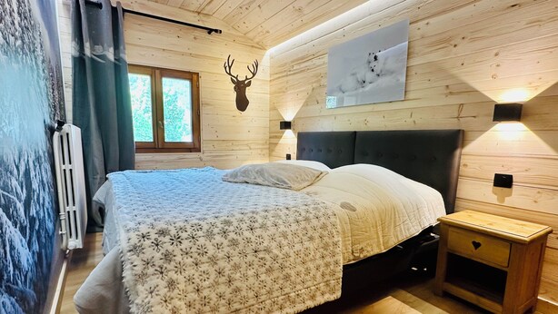 Un dormitorio de cabaña de madera con una cama, una cabeza de venado montada, una ventana y una lámpara fijada a la pared.