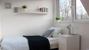 Une chambre bien rangée, avec un lit, une table de chevet, une lampe et une étagère où sont posés des livres et des plantes.