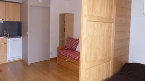 Een kleine kamer met een kitchenette, een bed, een houten garderobe en een inbouwplank.
