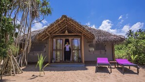 A beach hut with a thatched roof, a wooden door, and a small porch area. There are two lounge chairs with purple cushions and a small table.