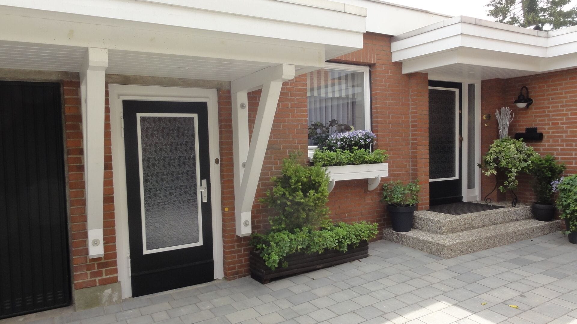 Een bakstenen huis met een zwarte deur, witte raamkozijnen en een klein terras met plantenpotten.