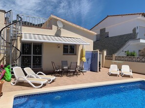 Un área junto a la piscina con un porche techado, sillones de descanso y una pequeña mesa.
