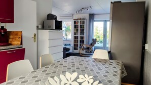 A compact kitchen with a microwave, a wooden cutting board, and a red appliance. A dining area with a patterned table and chairs is visible, along with a bookshelf and a window with a view of greenery.