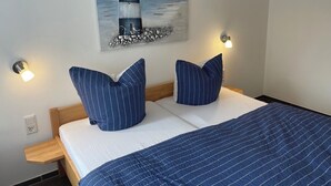 A neatly made bed with a striped blue bedspread and matching pillows, a painting of a lighthouse on the wall, and two wall-mounted reading lights. - FeWo Seestern... at home on vacation (Büsumer Deichhausen)